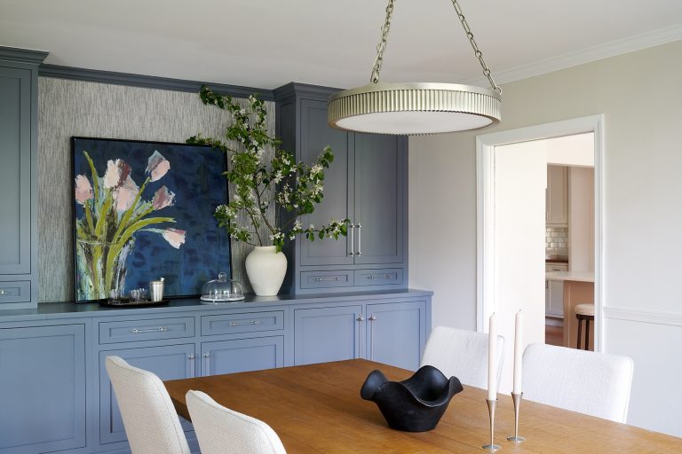 Designed by interior designer Glenna Stone, this dining room in Lafayette Hill features light blue cabinets, a large floral painting, and a wood table with cream chairs under a modern pendant light. A doorway leads to another room.