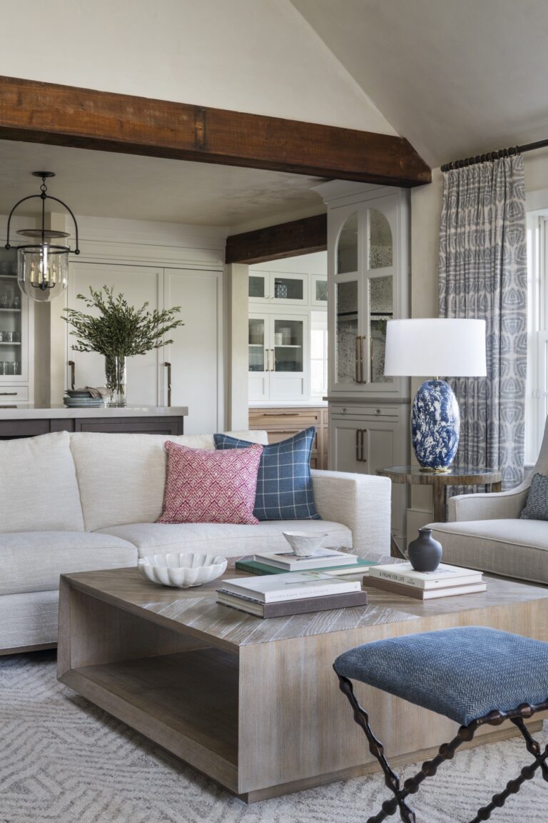 A cozy living room located in New Hope in Bucks County features a beige sectional with colorful pillows, a wood coffee table, and a blue-patterned lamp. Exposed beams and patterned curtains complete the inviting design by interior designer Glenna Stone.