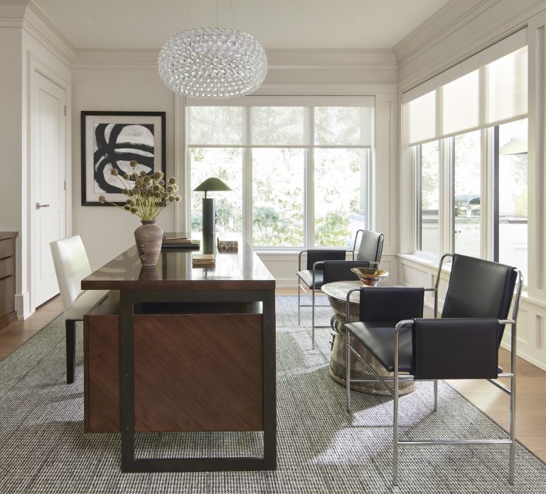 Modern office located on the Main Line with a wooden desk, two black chairs, and a coffee table on a gray rug. Large windows fill the space with light. Designed by interior designer Glenna Stone, it features an abstract painting and a crystal chandelier.