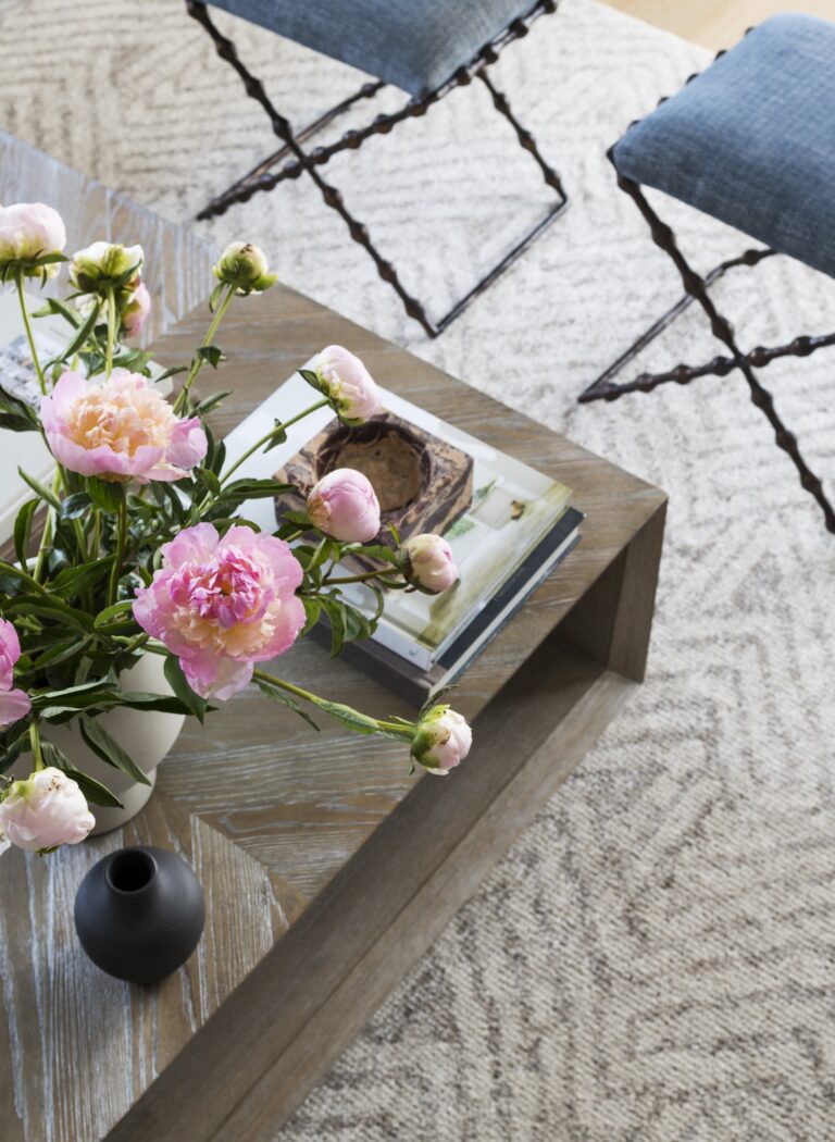 A wooden coffee table with a white vase holding pink peonies, a small black vase, a book, and a decorative object sits on a textured beige rug. Designed by interior designer Glenna Stone, it creates an inviting space located in New Hope in Bucks County.