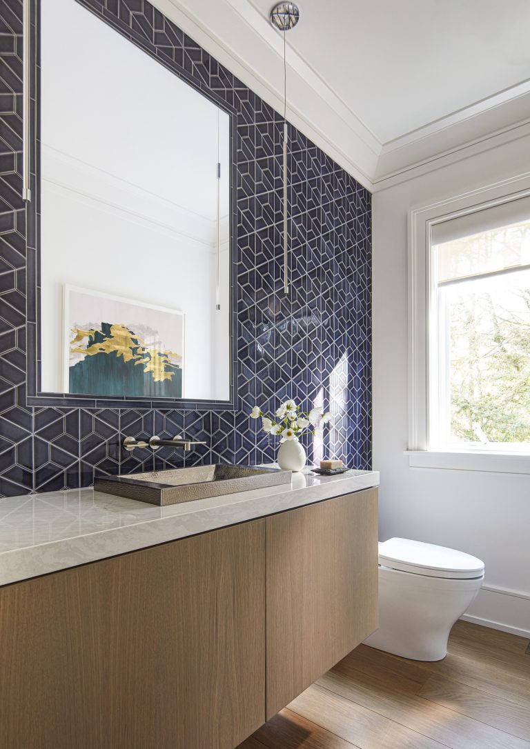 A modern bathroom located on the Main Line features a dark geometric hexagon tile wall, floating wood vanity with marble countertop, and framed art—styled by interior designer Glenna Stone to maximize natural light and elegant simplicity.