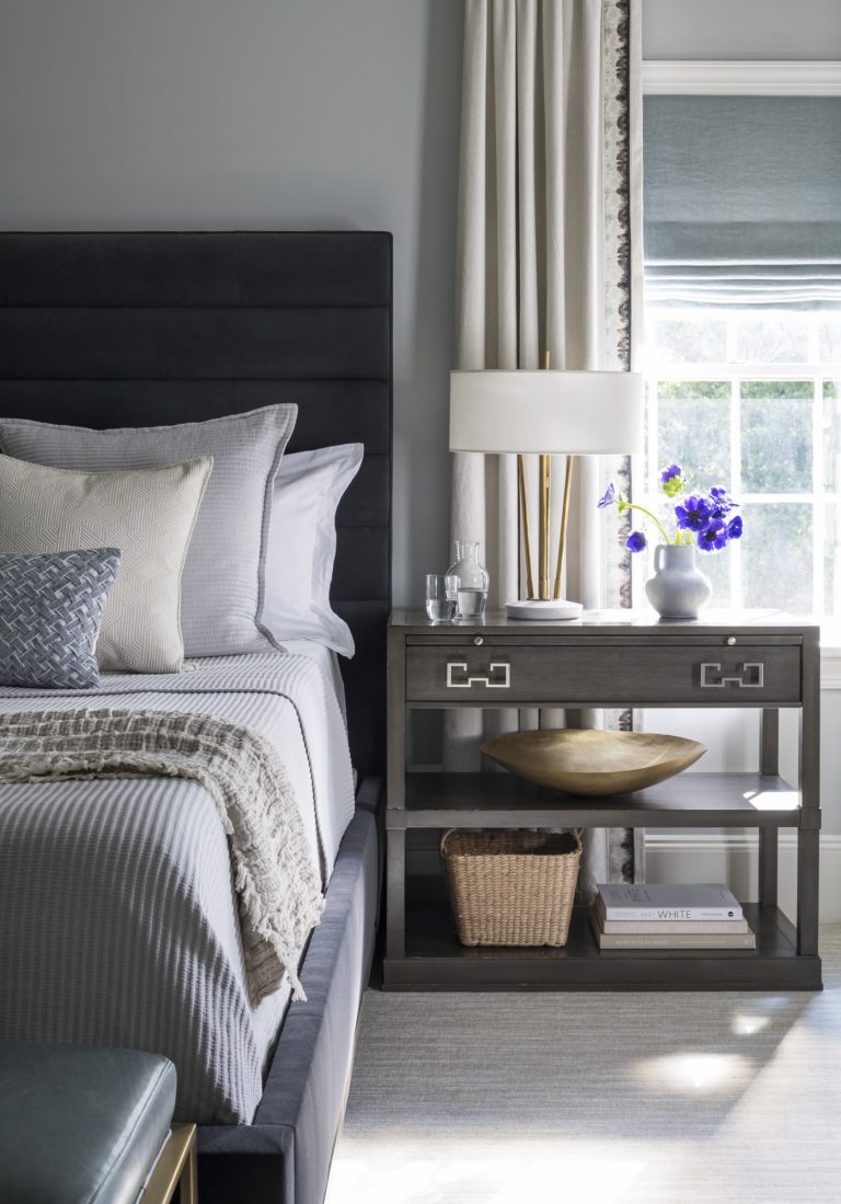 A stylish bedroom, designed by interior designer Glenna Stone, features a neatly made bed with gray and white bedding, a padded headboard, and layered pillows. Sunlight filters in through white curtains and a blue shade in this Main Line oasis.