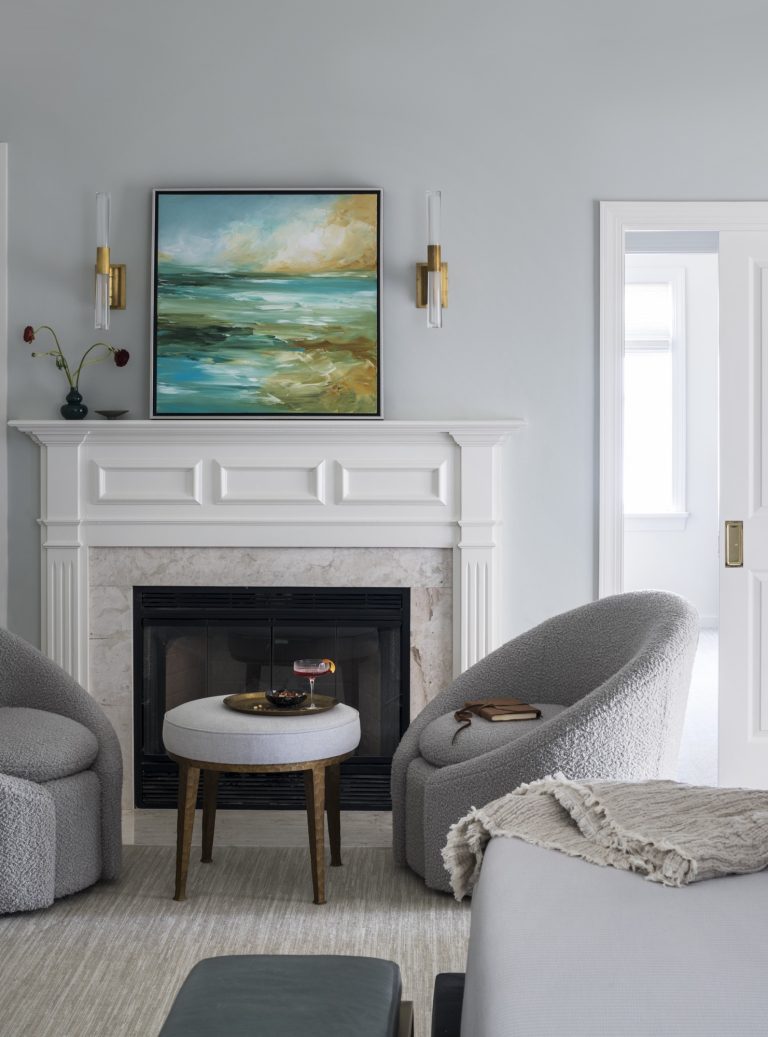A modern living room by interior designer Glenna Stone, located in Gladwyne on the Main Line, features curved gray chairs, a round ottoman table, abstract seascape art, gold sconces, and a cozy blanket on the bed in the foreground.