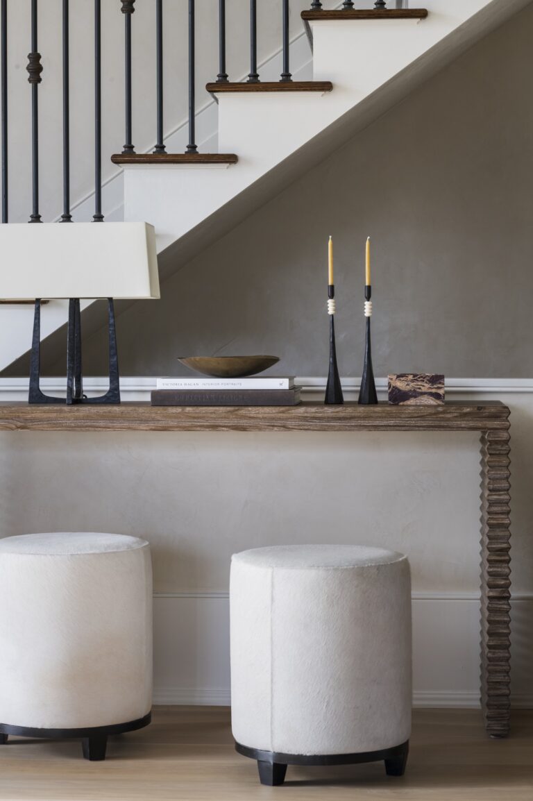 A modern entryway designed by interior designer Glenna Stone features a rustic wooden console table, white cylindrical stools, black candlesticks with yellow candles, books, a bowl, and a lamp. The space is located in New Hope in Bucks County.