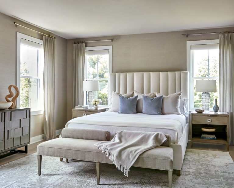 A modern, airy bedroom by interior designer Glenna Stone features a tall, tufted beige headboard, neutral bedding with blue accent pillows, and is located in Bucks County. Sheer curtains frame the windows beside each stylish nightstand.
