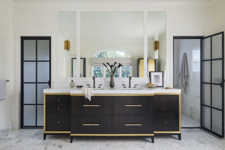 Modern bathroom located in Gladwyne on the Main Line, featuring a black double-sink vanity with gold accents, black-framed mirrors, marble floor, decorative items, frosted glass doors, and a large window reflecting greenery. Interior designer Glenna Stone.