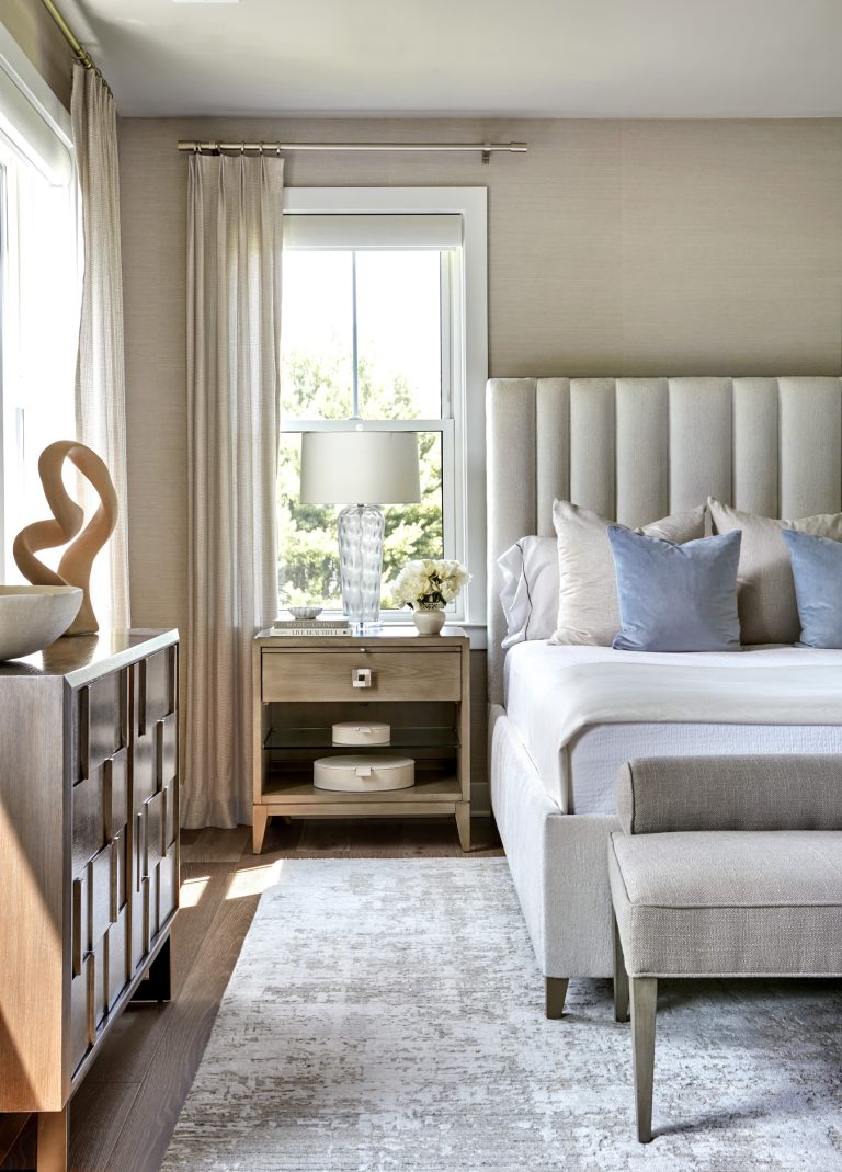 A bright, modern bedroom by interior designer Glenna Stone, located in Bucks County, features a tall upholstered headboard, a bed with white and light blue pillows, a textured rug, and a wood dresser topped with sculptural decor.