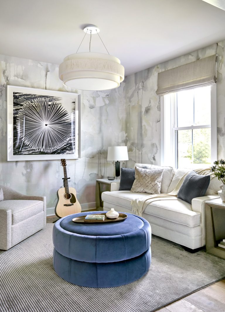 Modern living room by interior designer Glenna Stone, located in Bucks County, featuring abstract wallpaper, a white sectional with gray and cream pillows, blue velvet ottoman, gray armchair, acoustic guitar, and a sunlit window with Roman shade.
