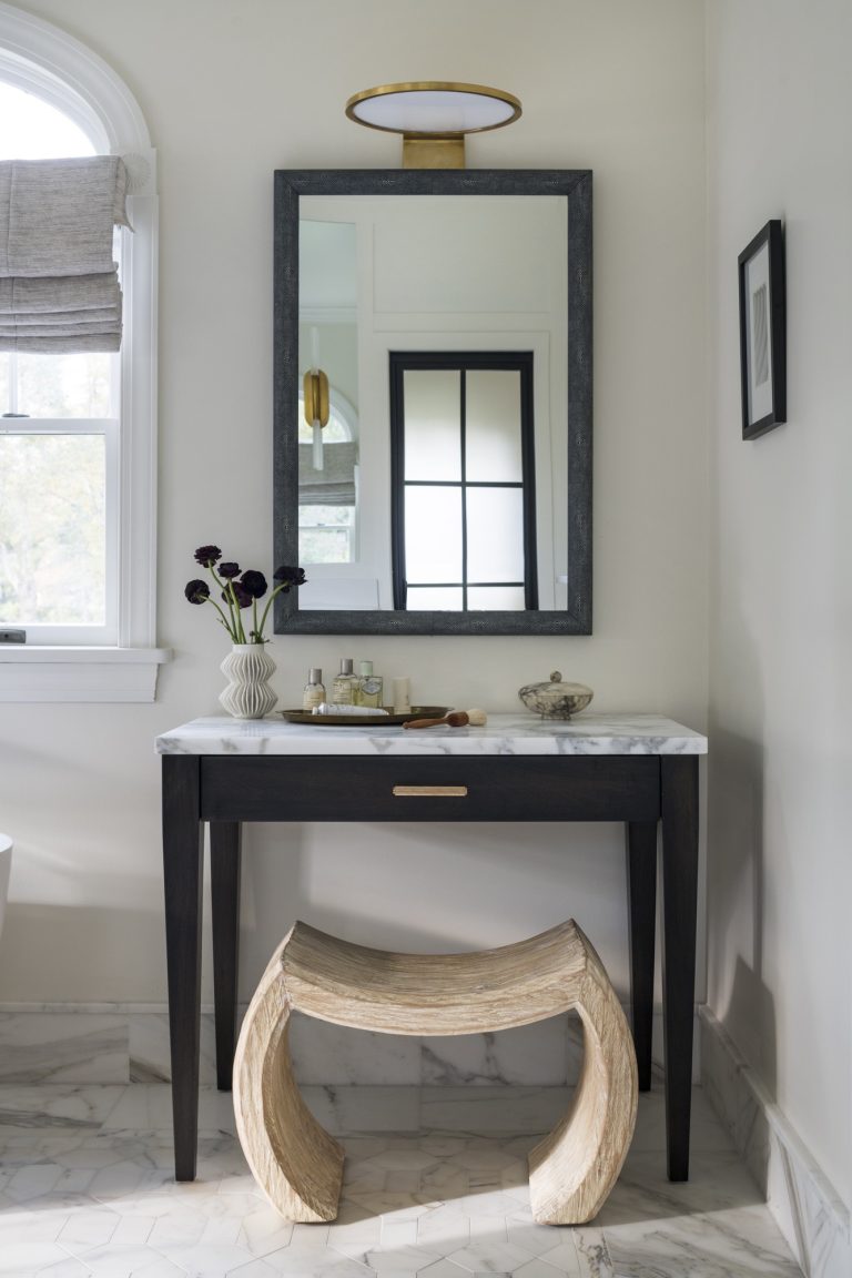 A bathroom vanity, designed by interior designer Glenna Stone, features a black-framed mirror, marble countertop, and a modern curved wooden stool. Located in Gladwyne on the Main Line, the space boasts light walls, marble floors, and a Roman shade.