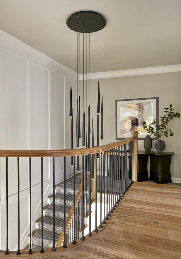 Modern staircase landing by interior designer Glenna Stone, located in Bucks County, with a curved wooden handrail, black vertical balusters, and grey carpeted stairs. A black pendant chandelier hangs above a console table with vases and abstract art on the beige wall.
