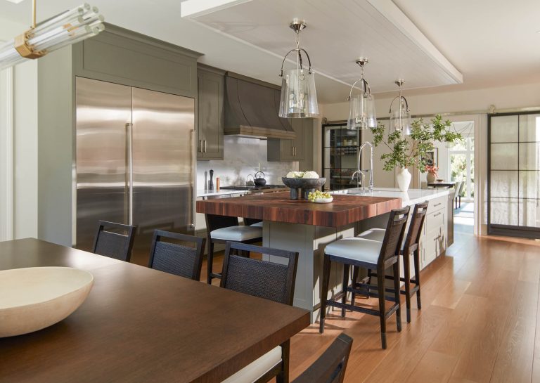 Modern kitchen located on the Main Line with wood flooring, a large island featuring a wood countertop, bar stools, pendant lights, stainless steel appliances, green cabinetry, and curated by interior designer Glenna Stone. Natural light fills the space.