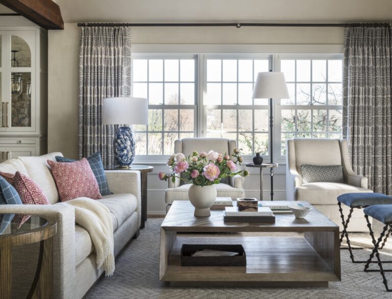 A bright living room designed by interior designer Glenna Stone features a light sofa, patterned cushions, and a cozy throw. Two armchairs face a wooden coffee table with pink flowers. Large windows with patterned curtains fill this New Hope, Bucks County home with sunlight.