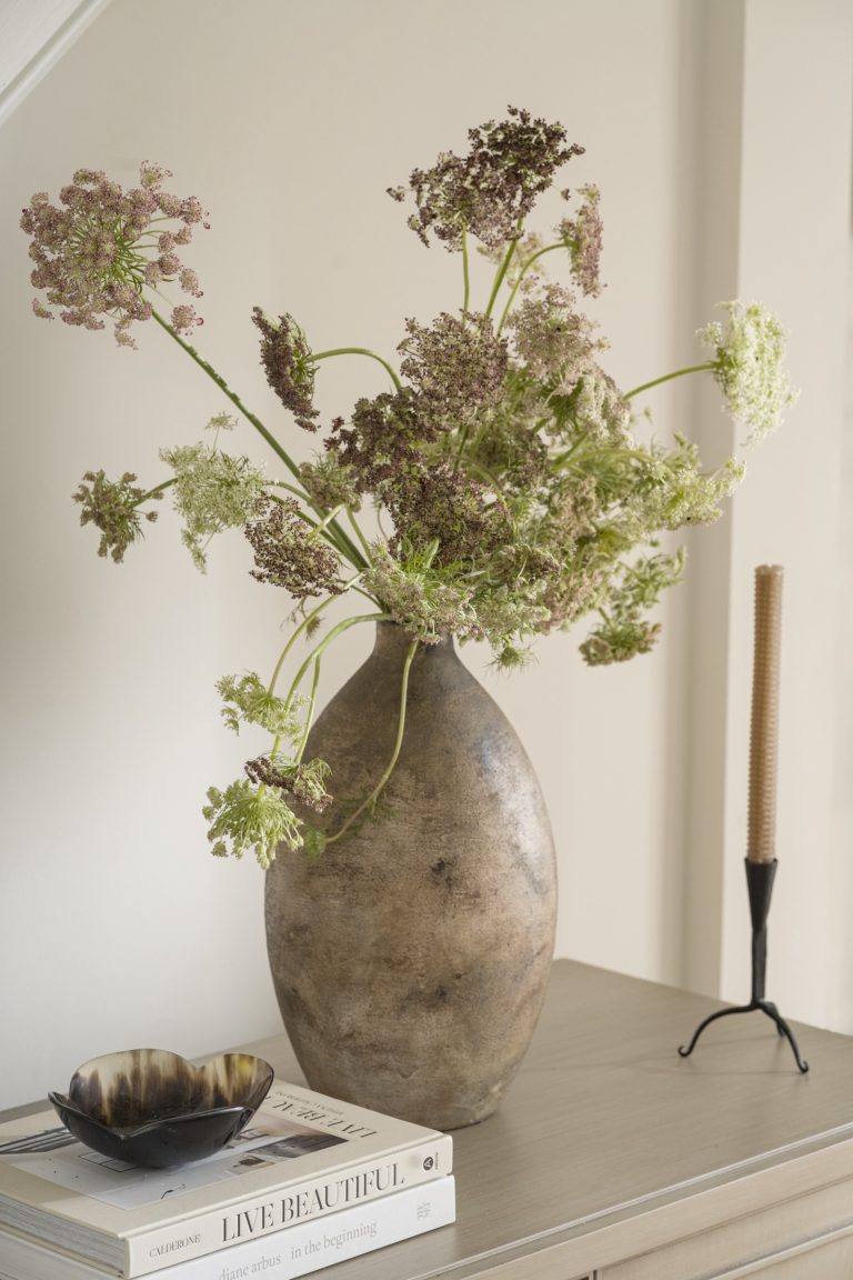 A rustic brown vase with tall, wispy Queen Anne’s Lace flowers sits on a beige table. Curated by Main Line decorator Glenna Stone and located in Penn Valley, this serene scene features stacked books and a spiral candle for natural elegance.