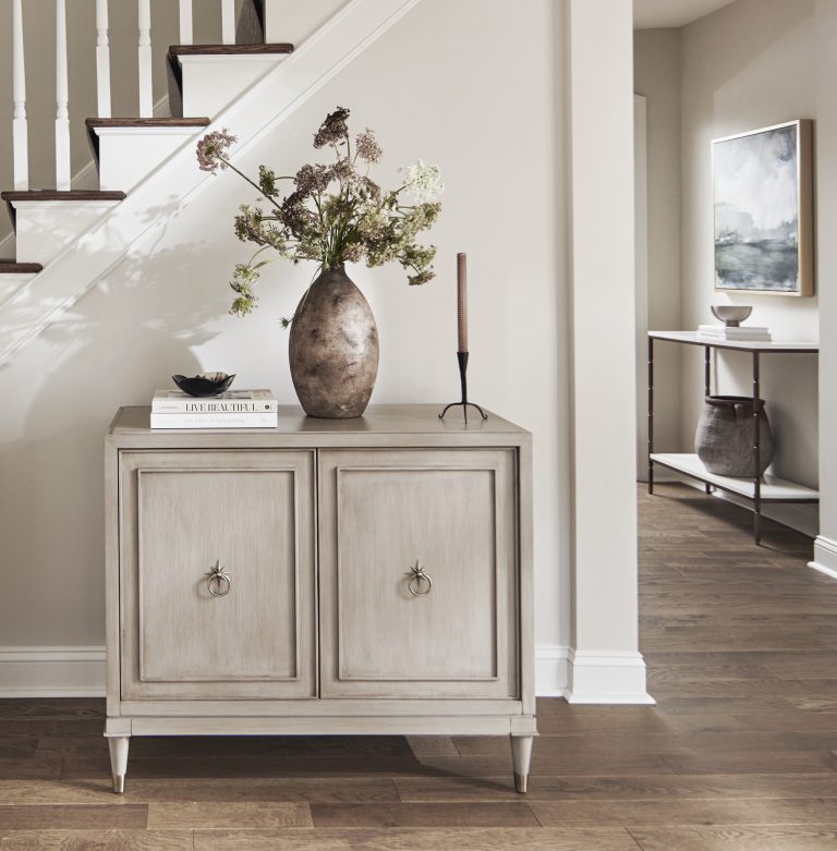 A light wood cabinet with two doors stands against a white wall below a staircase. Styled by Main Line decorator Glenna Stone, it’s located in Penn Valley and topped with a rustic vase of flowers, a book, and a tall candlestick.