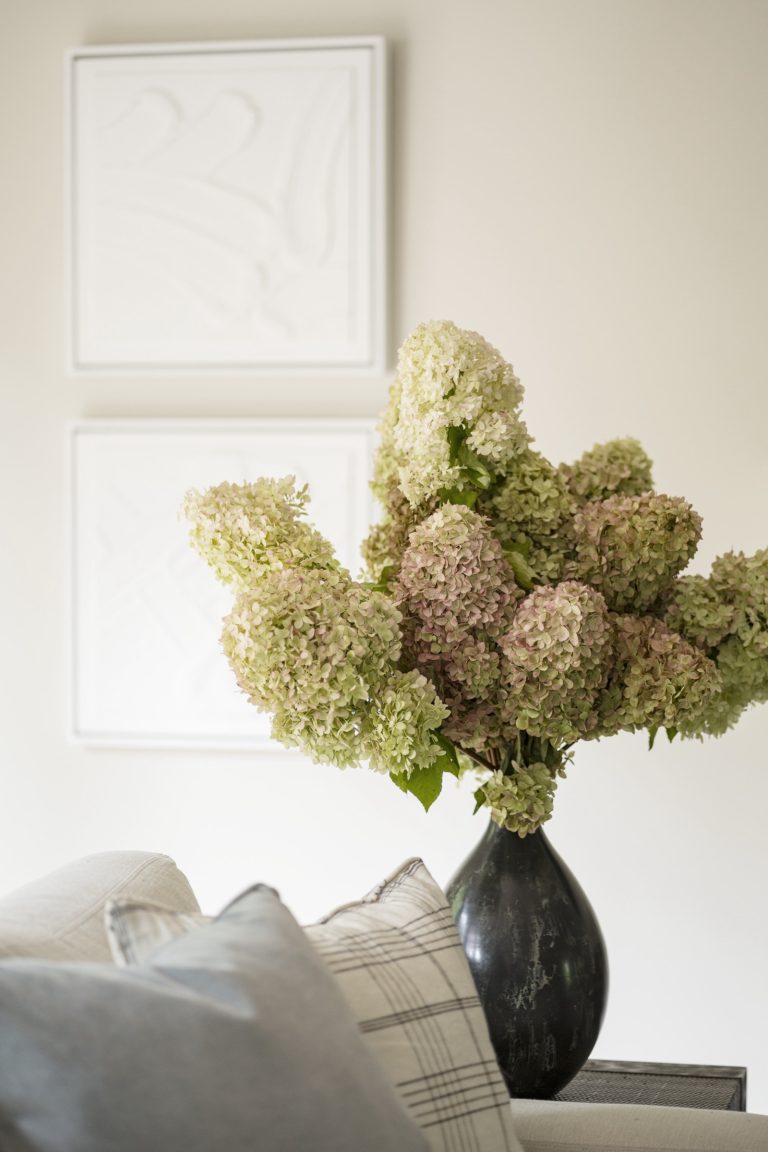 A dark vase filled with pale green and pink hydrangeas sits on a table behind a sofa with plaid and grey pillows; two white, textured square artworks hang on a light-colored wall in this space by Main Line decorator Glenna Stone, located in Penn Valley.