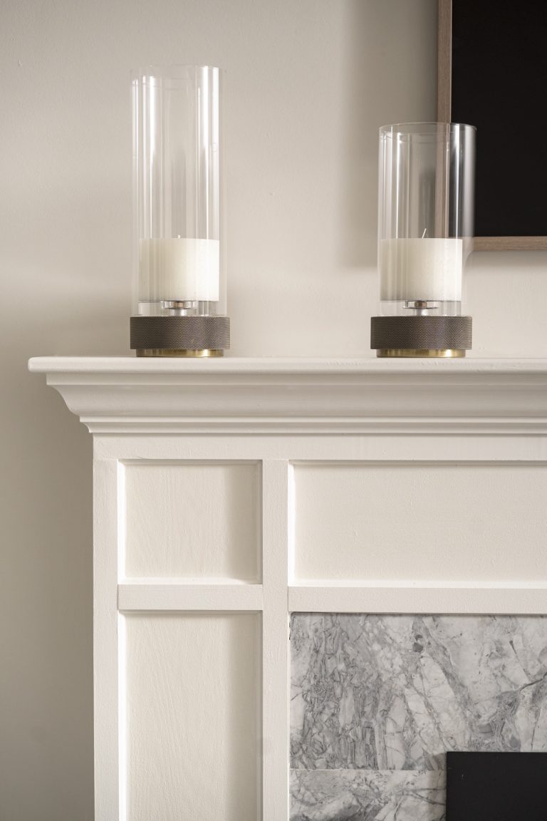 Located in Penn Valley, this white mantel with decorative trim—styled by Main Line decorator Glenna Stone—holds two cylindrical glass hurricane candle holders. Below, light gray marble with darker veining frames the elegant fireplace.