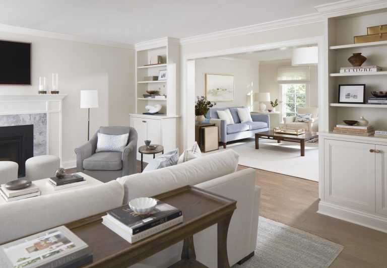 A bright, modern living room located in Penn Valley features white walls, built-in shelves, neutral-toned furniture, a gray armchair, and a fireplace. Designed by Main Line decorator Glenna Stone, the adjoining room offers an airy and inviting vibe.