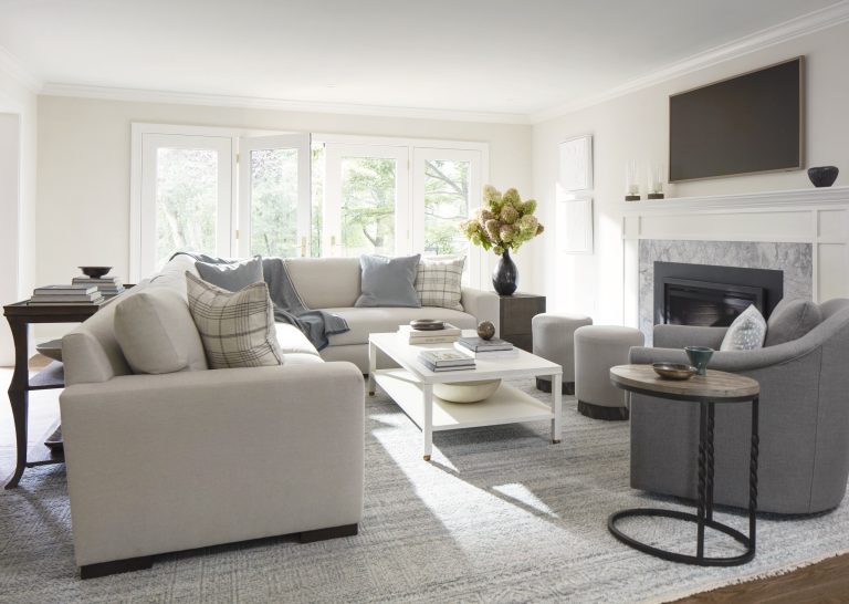 A bright, modern living room located in Penn Valley features light gray sofas, a white coffee table, two round ottomans, and a fireplace with a flat-screen TV above it. Minimal, elegant decor reflects the touch of Main Line decorator Glenna Stone.