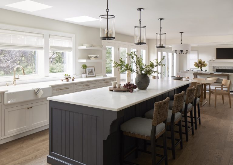 A spacious, modern kitchen by Main Line decorator Glenna Stone, located in Penn Valley, features a large dark gray island, white countertops, wicker chairs, and pendant lights. White cabinets and greenery in a vase brighten the airy space.