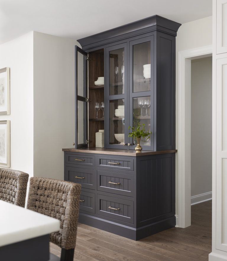 A dark gray built-in china cabinet by Main Line decorator Glenna Stone displays white dishes and glassware. Located in Penn Valley, the space features gold hardware, a wooden countertop with greenery, woven dining chairs, and framed wall art.