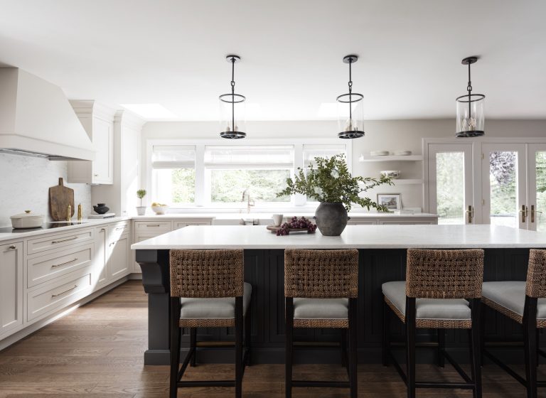 Modern kitchen in Penn Valley designed by Main Line decorator Glenna Stone, featuring white cabinets, a large black island with a white countertop, woven barstools, pendant lights, and wood flooring. Big windows fill the space with natural light.