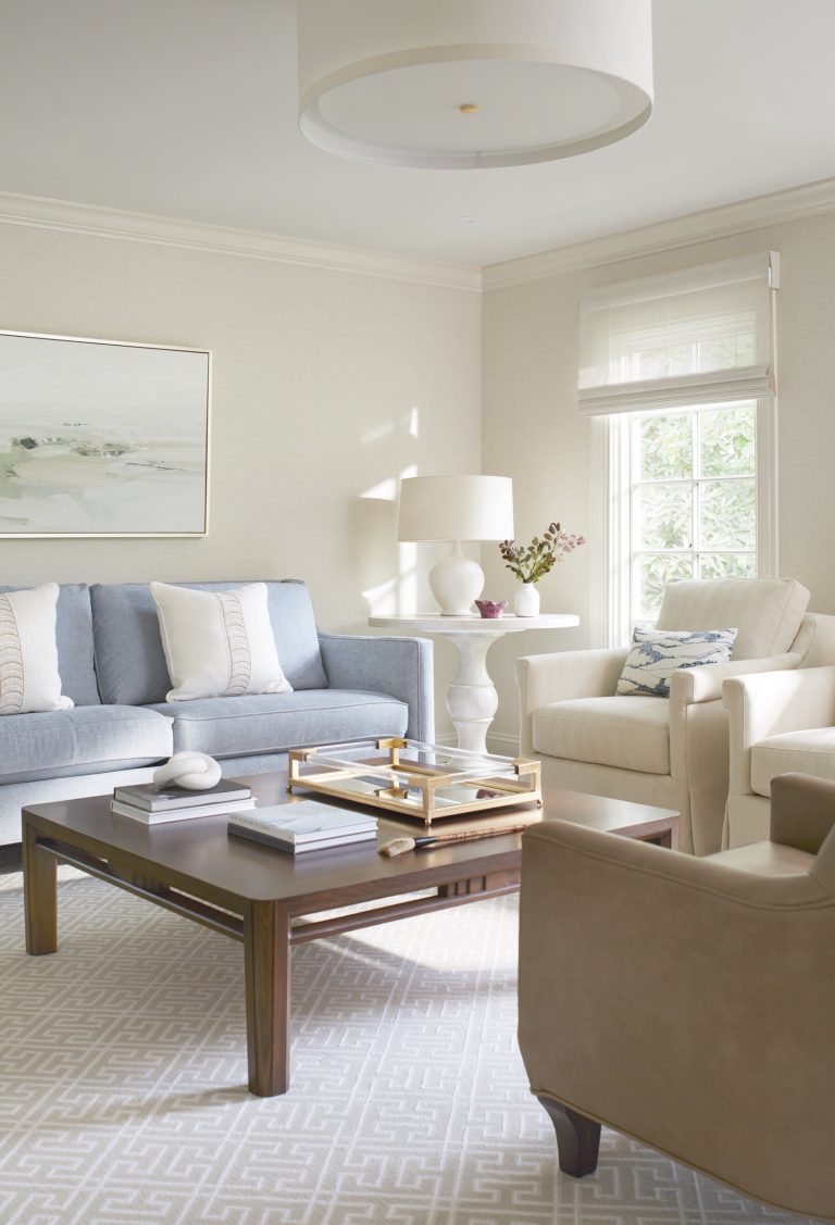 Bright living room by Main Line decorator Glenna Stone, located in Penn Valley, features a pale blue sofa, cream armchairs, and dark wood coffee table. Light streams through sheer blinds, creating a calm, airy feel accented by flowers and decorative books.