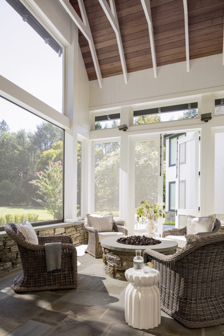 A bright sunroom, designed by interior designer Glenna Stone, features vaulted ceilings and exposed beams with wicker armchairs around a fire pit table. Large windows frame lush Main Line views, filling the airy space with natural light.