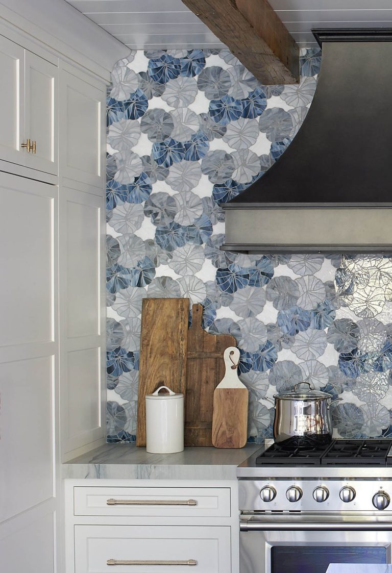 A modern kitchen corner designed by interior designer Glenna Stone, located in New Hope in Bucks County, features a gas stove, metallic range hood, floral tile backsplash, wooden cutting boards, and sleek white cabinets on the left.
