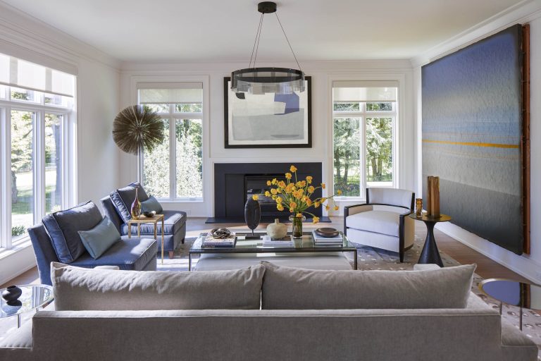 A bright, modern living room located on the Main Line features large windows, a gray sofa, blue armchairs, a white accent chair, a glass coffee table with yellow flowers, abstract art, and contemporary décor by interior designer Glenna Stone.