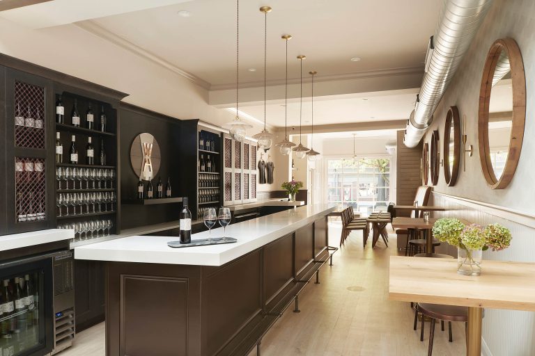 A modern, bright wine bar in Haddonfield with a long white counter, dark cabinets, wine bottles and glasses on display, hanging pendant lights, round mirrors on the right wall, wood tables with chairs, large windows, and green hydrangeas in a vase.