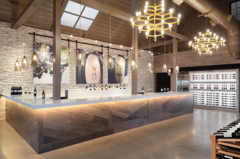 Modern winery tasting room in Philadelphia, designed by interior designer Glenna Stone for William Heritage Winery, features a large angular bar, soft backlighting, striking chandeliers, and wine-themed decor in a stunning stone-and-wood accented space.