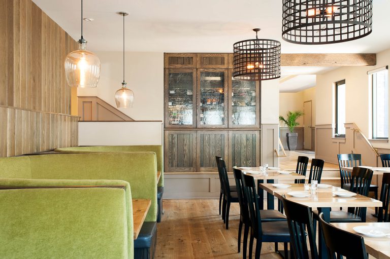 Modern restaurant interior by interior designer Glenna Stone, featuring green upholstered booth seating, black wooden chairs, and light wooden tables set with white plates. Pendant lights illuminate wood paneling and glass cabinets as daylight streams in.