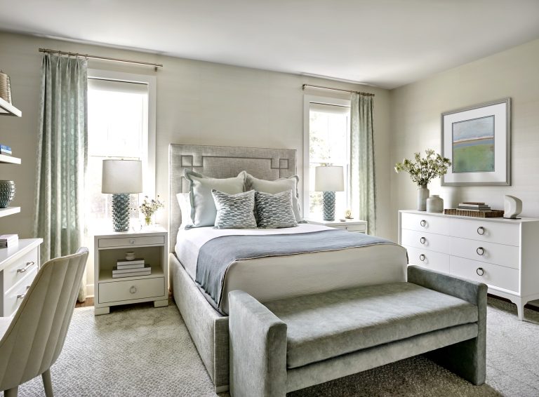 A modern bedroom located in Bucks County features a gray upholstered bed, silver bench, and matching pillows. Designed by interior designer Glenna Stone, the room includes sheer curtains, chic art, white nightstands, and a stylish desk with chair.