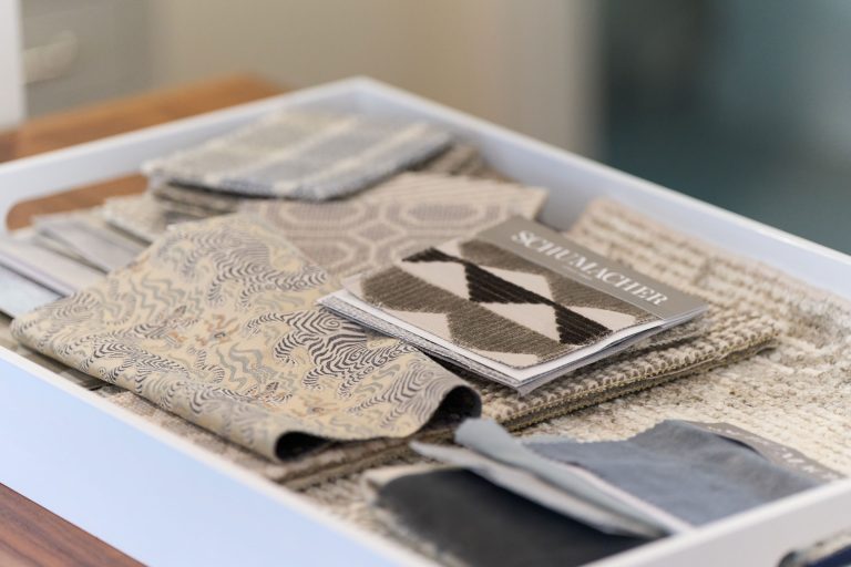 A white tray holds neatly stacked fabric swatches in various neutral tones and patterns, including geometric, abstract, and textured designs. A SCHUMACHER booklet rests on top—a refined vignette inspired by Glenna Stone Interior Design. The background is softly blurred.