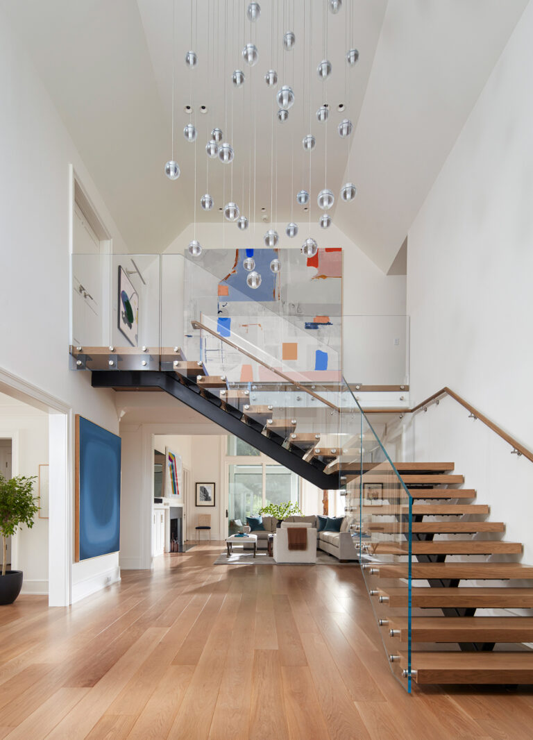 A modern open-concept living space located on the Main Line, featuring a floating wooden staircase with glass railing, abstract art, wood floors, contemporary furniture, lush greenery, and striking silver pendant lights from the high white ceiling.
