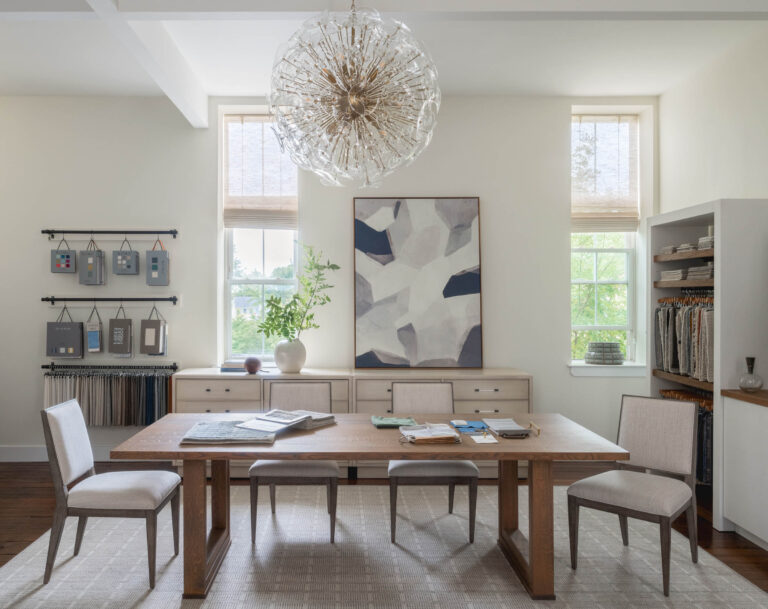 A modern dining room located on the Main Line features a wooden table, four beige chairs, a large abstract painting, and a white sideboard. Designed by the office of Glenna Stone Interior Design, it boasts bookshelves, fabric samples, and a spherical chandelier.