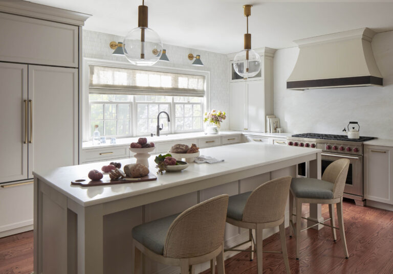 A bright, modern kitchen located in Bryn Mawr features light cabinetry, a spacious island with three cushioned stools, and stylish pendant lights. Interior designer Glenna Stone’s touch is seen in the fresh florals by the sink and elegant Roman shade.