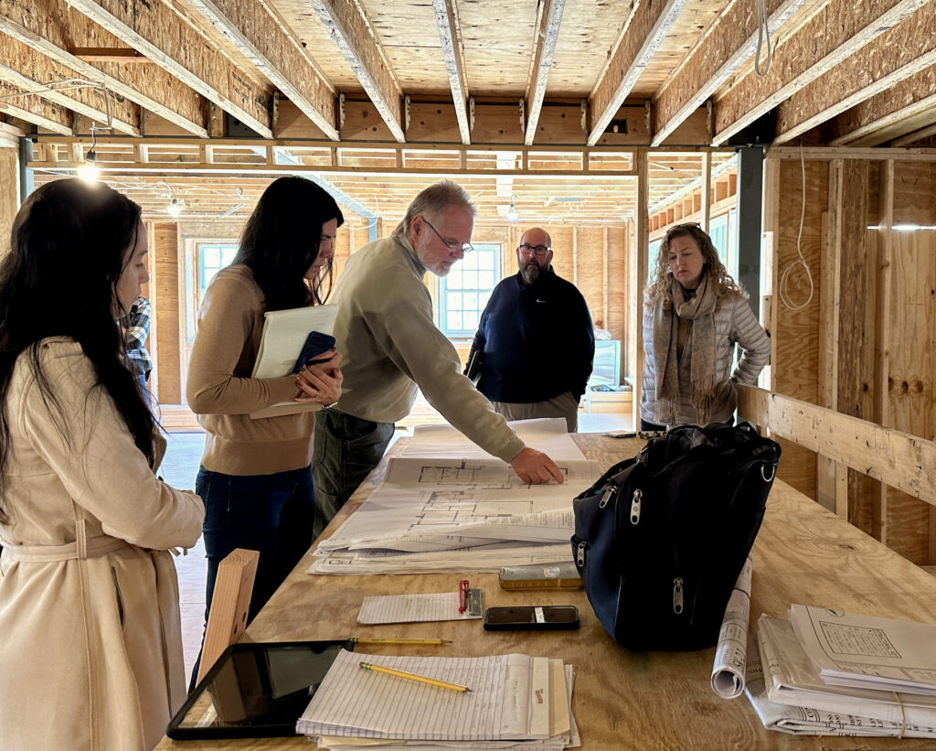 Five people, including Glenna Stone Interior Design team members, stand around a table with blueprints in a house under construction. One man points at the plans while others observe and take notes amid exposed beams and unfinished walls.