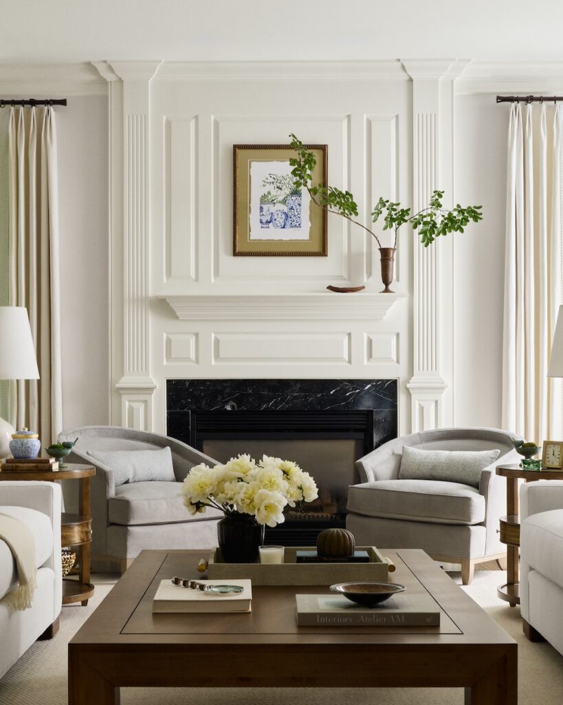 Elegant living room with white walls, black marble hearth, two gray armchairs, and a wooden coffee table styled with books and flowers.
